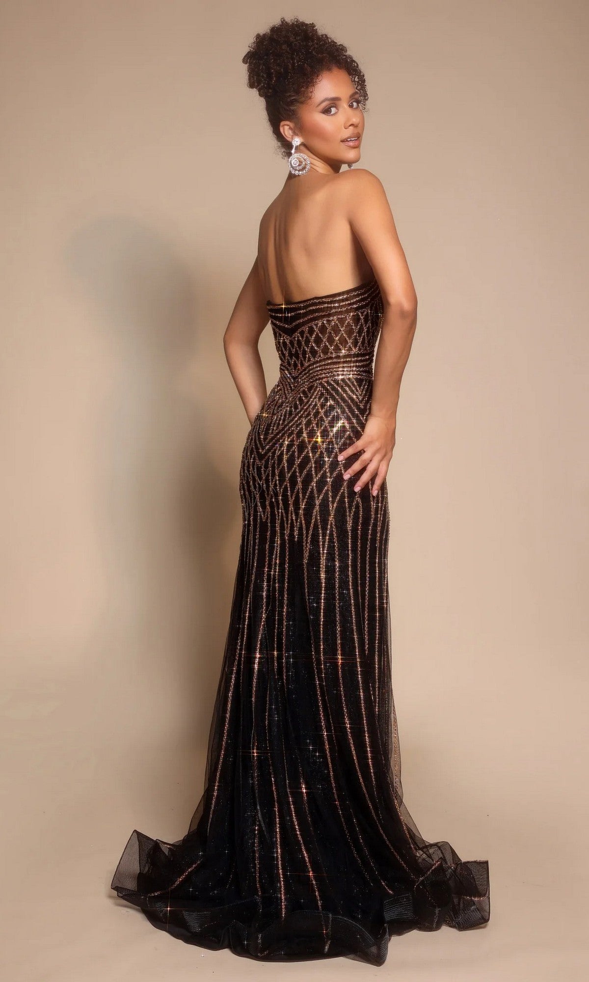 Model poses against a plain beige background, looking over her shoulder in the Portia and Scarlett PS26072—a strapless long formal dress featuring geometric gold embellishments and a thigh-high slit.
